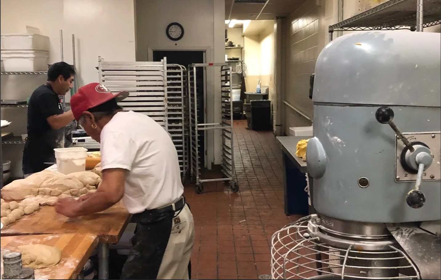 Dos panaderos trabajan masa junto a charolas y una batidora grande en la cocina de producción.