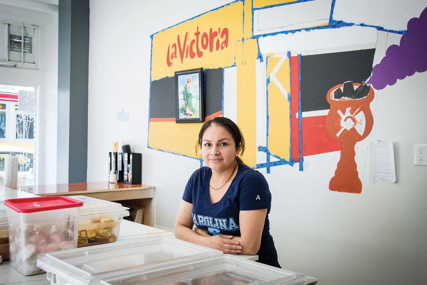 Una trabajadora de la panadería está sentada en el mostrador frente a un mural pintado de La Victoria.
