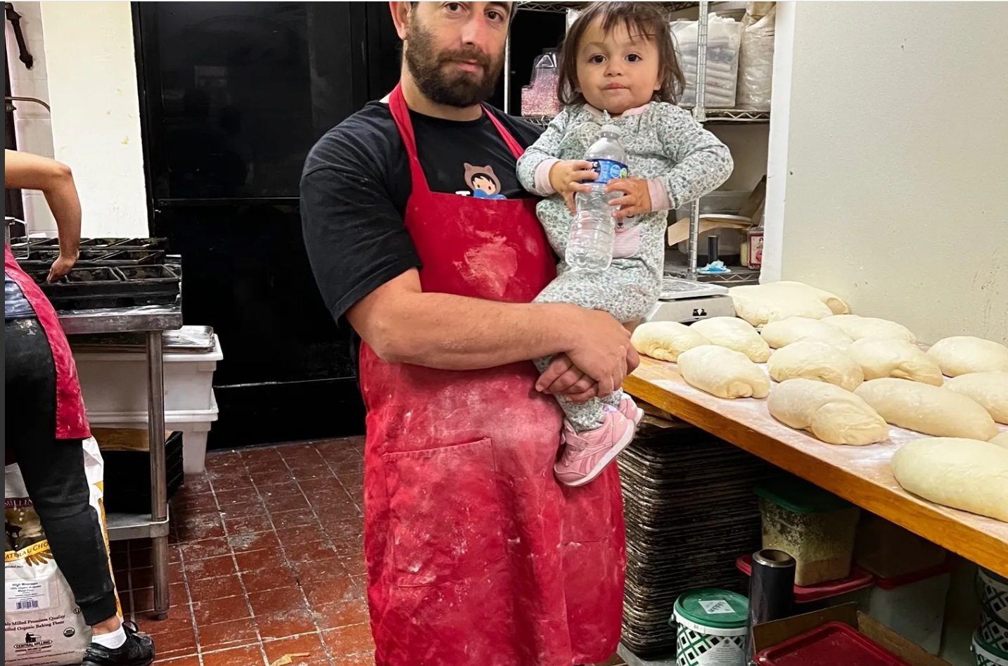 Un panadero sostiene a una niña pequeña junto a una mesa cubierta de masa formada.